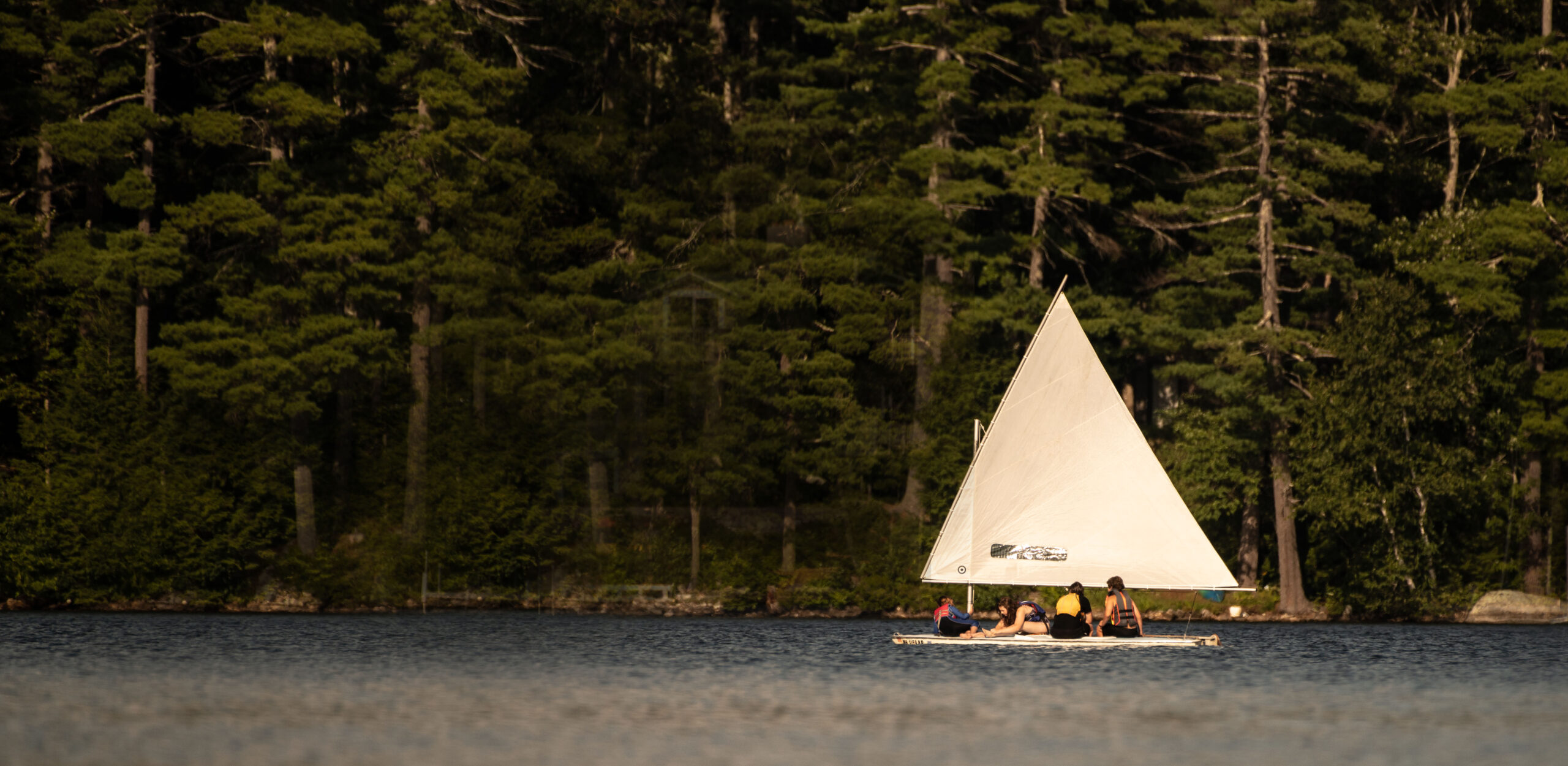 A Sunfish sailboat with four kids on it sailing on Lovell Lake.